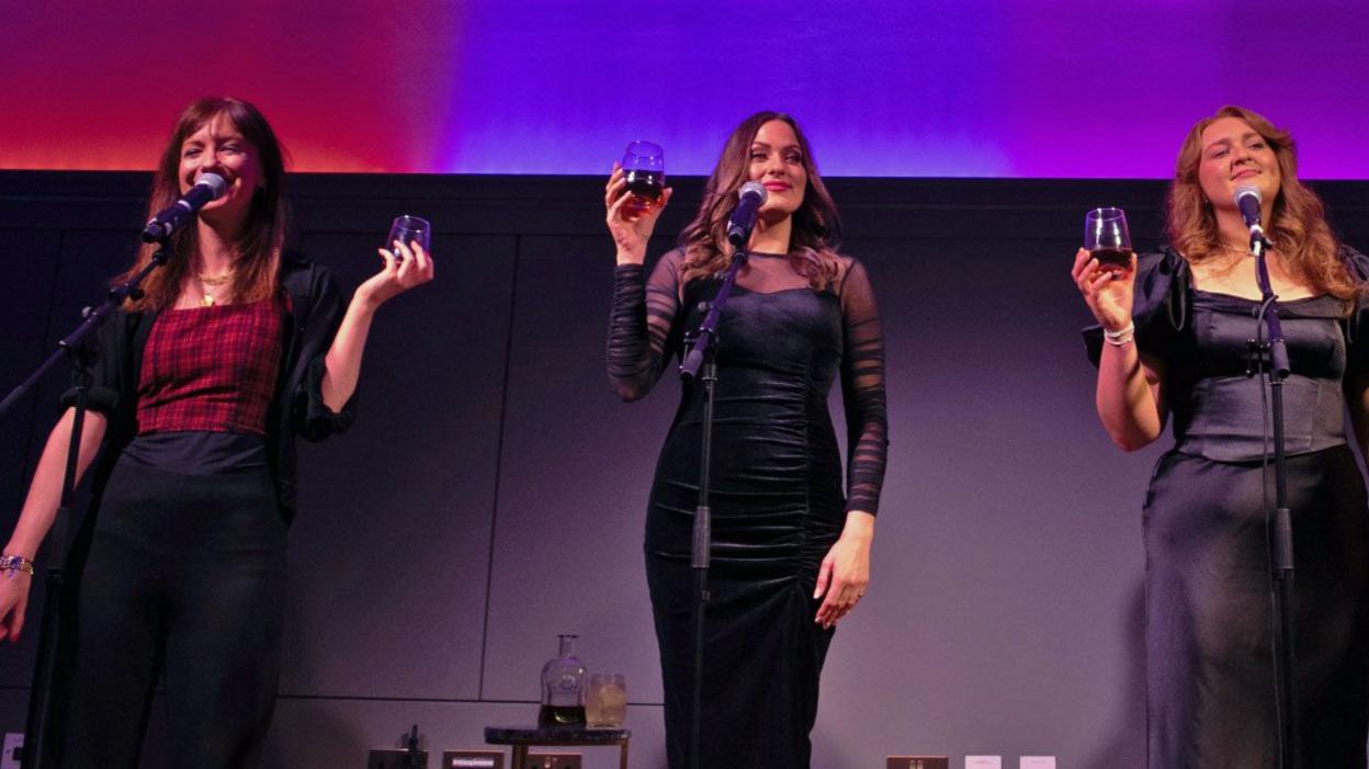 Three women stand together, raising a toast. They are elegantly dressed nd stand in front of a purple and pink backdrop.