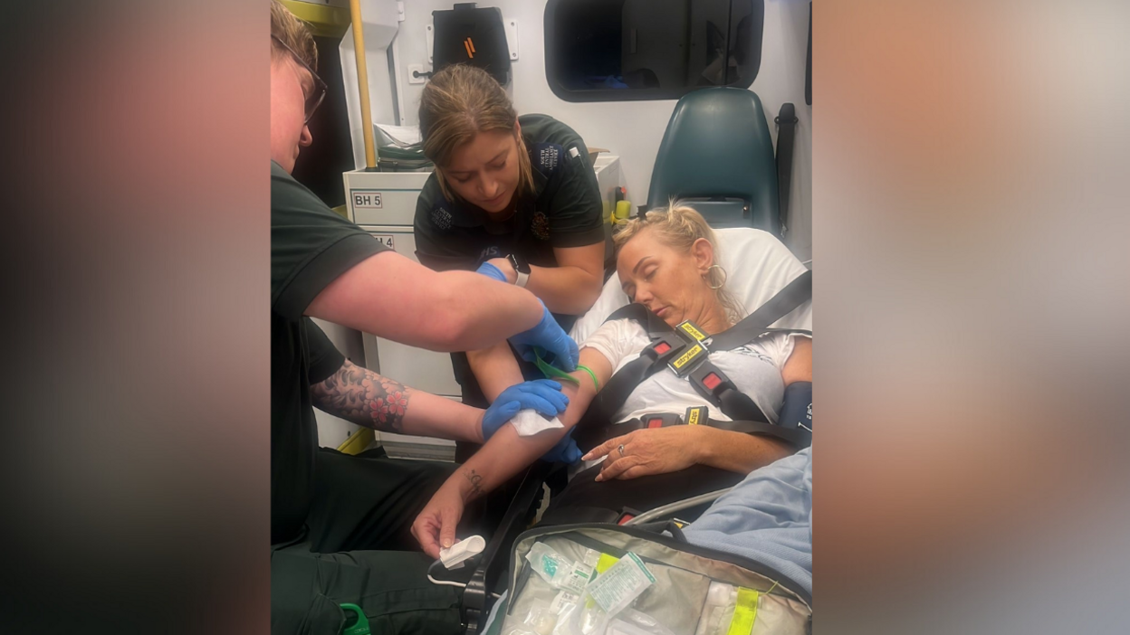 Michelle Sword being helped by paramedics. She lies unconscious, strapped to an ambulance bed. A man and a woman paramedics are assisting her.