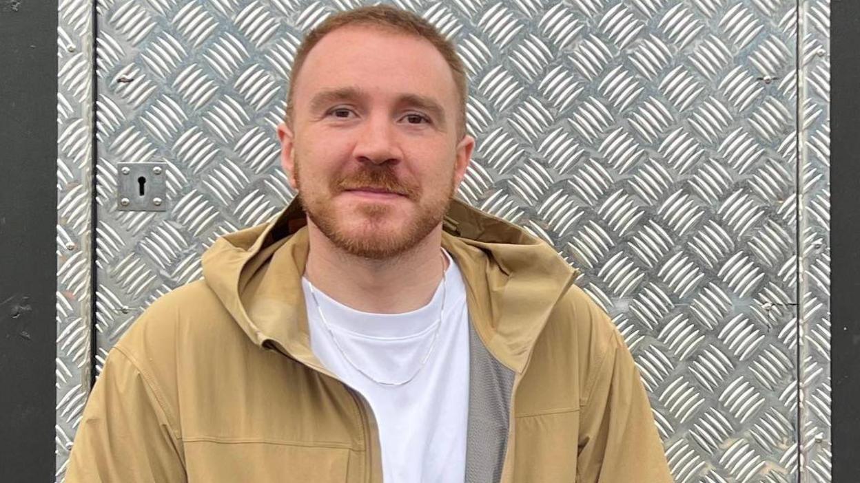 Chris Hindle stands outside his XLR club in Manchester. He is wearing a white t-shirt and light brown jacket. He has short, light brown hair and stubble.