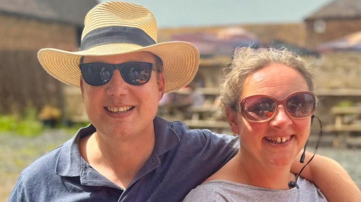 A man wearing a hat, sunglasses and a dark blue shirt has his arm around a woman wearing a grey shirt and sunglasses. Both are smiling at the camera and standing in a courtyard at a farm.