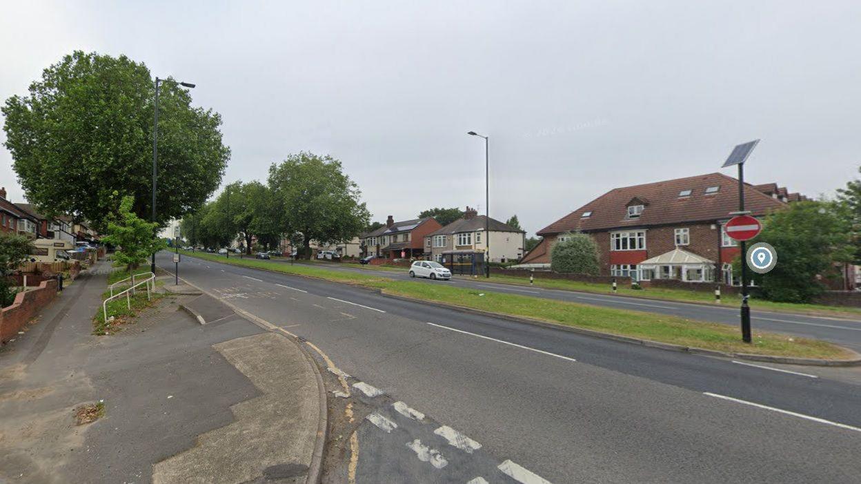 A street view of the road which is situated in a residential area with several houses along either side of the street. A white car can be seen driving down the road.