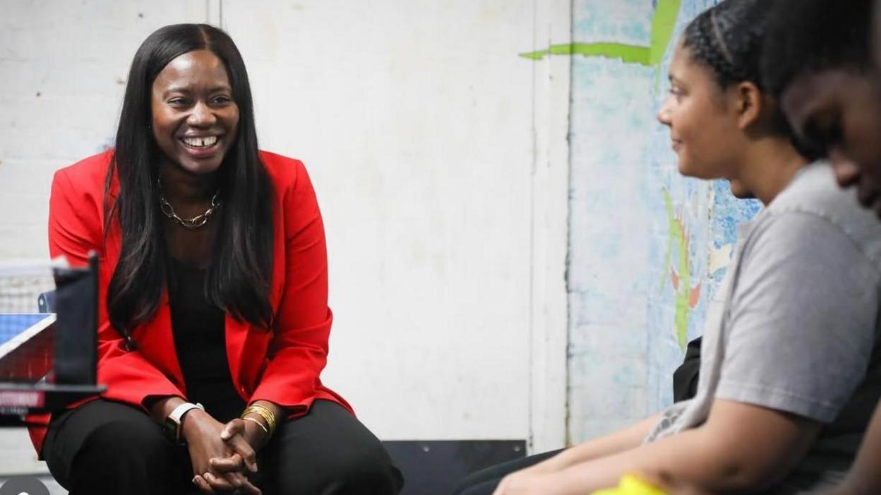 A woman in a bright red jacket and black trousers and top smiles as she speaks to some young people at a youth centre.