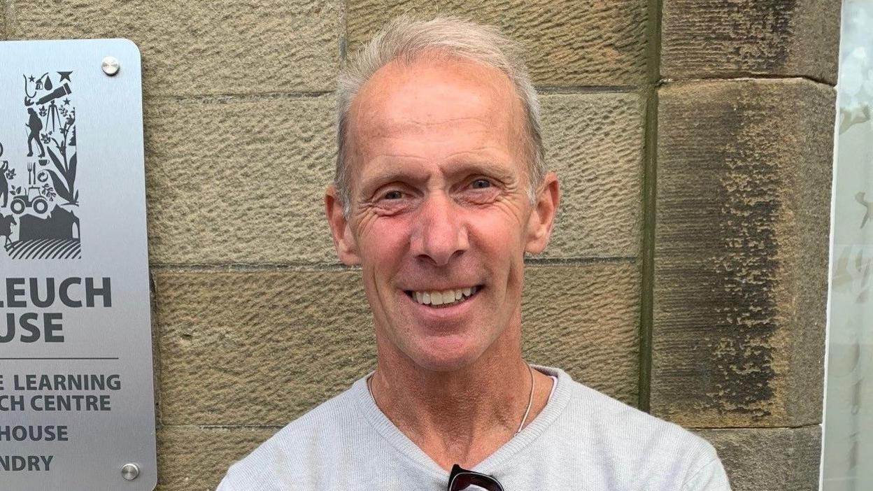 A man with thinning grey hair in a grey jumper with sunglasses hanging from the neck standing in front of a granite wall
