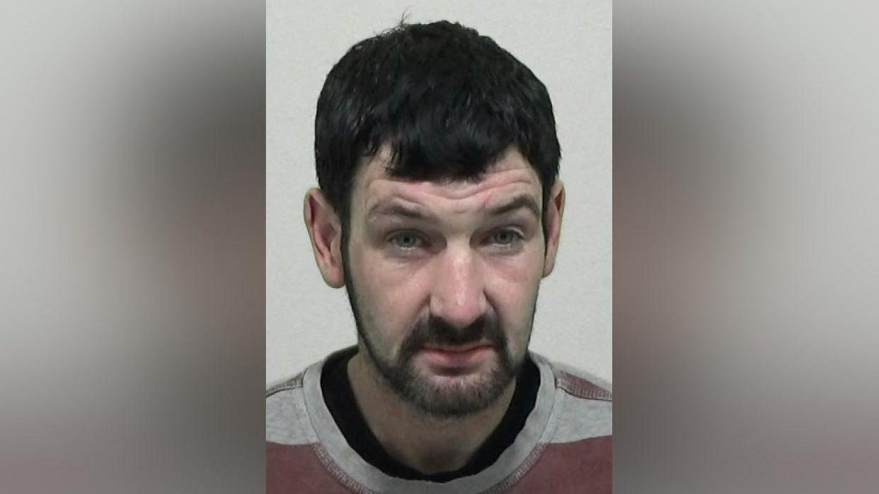 Mugshot of Layton Wood. He has short thick black hair and a black beard, with a dazed expression on his face.