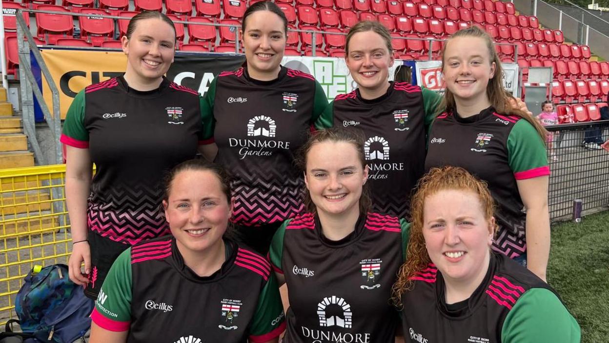 Seven women pose in a football team style photograph. They are wearing matching kits, black shirts with green sleeves. The words Dunmore Gardens are on the front of the shirts. All the women are smiling.