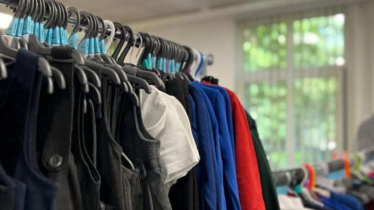 A rail of uniforms with white shirts, blue jumpers, grey pinafores and red sweatshirts on hangers.