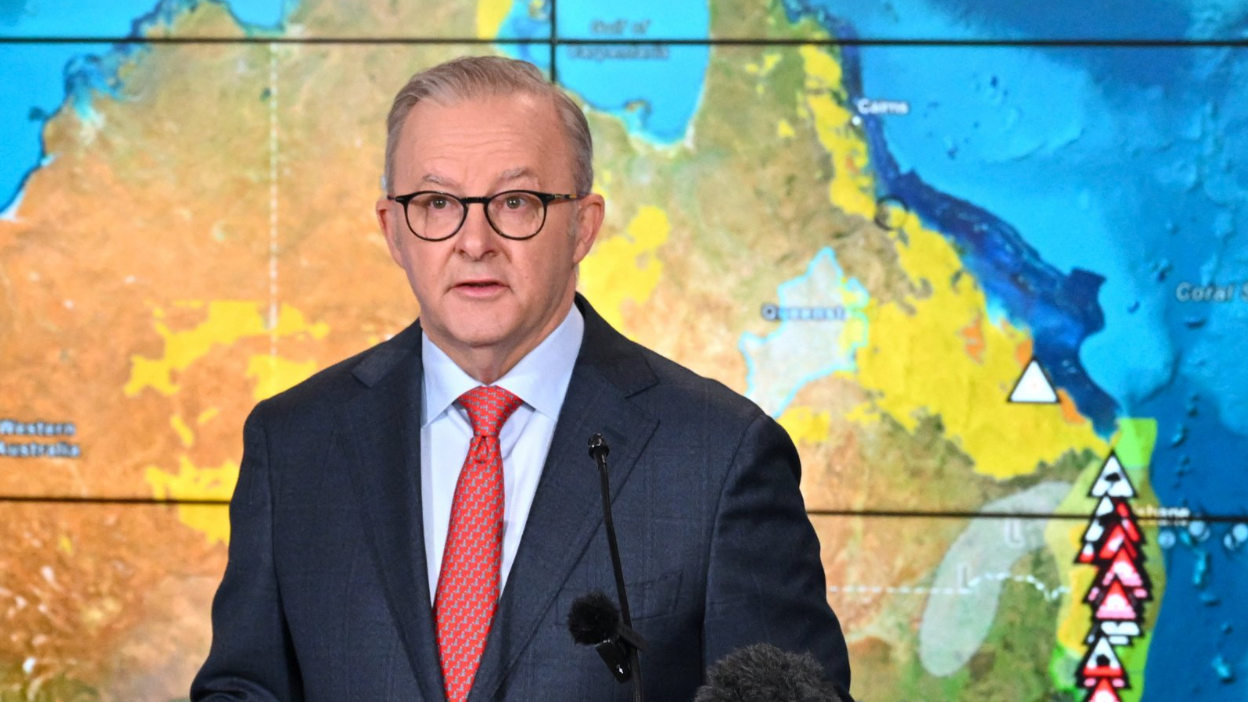 The Australian prime minister delivering a briefing in front of a screen showing weather warnings for the country's east coast