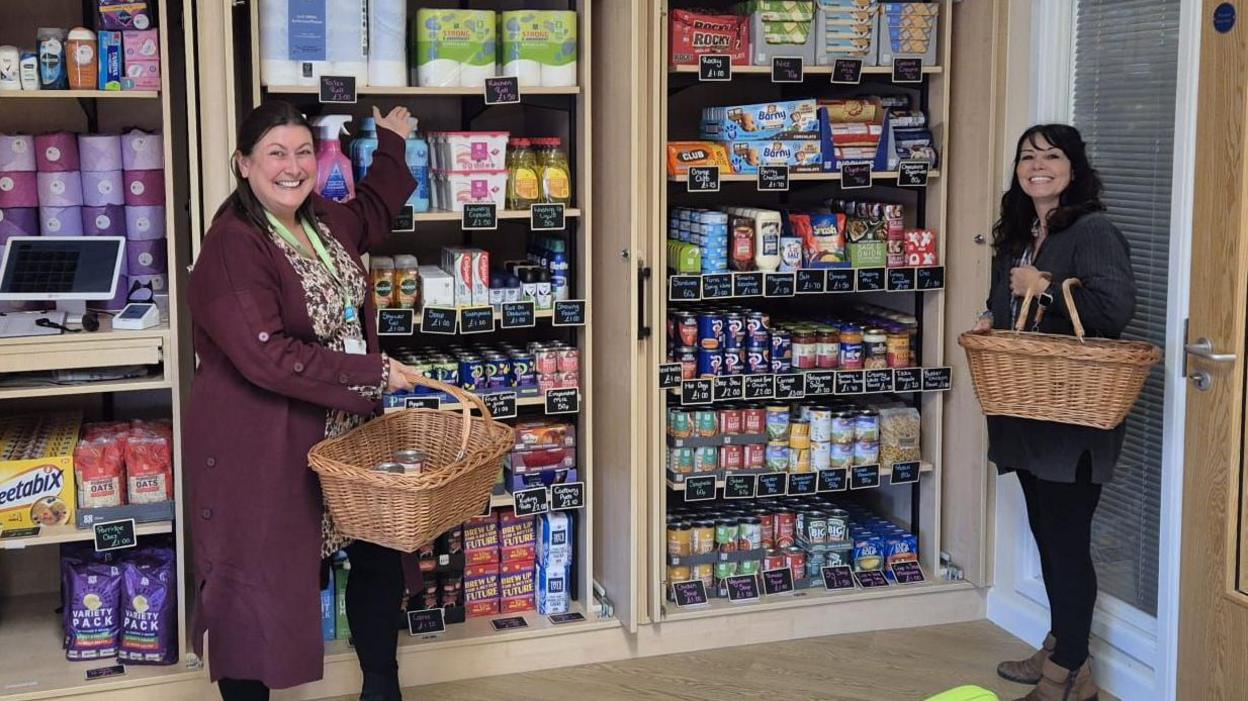 A pair of women holding wicker baskets stand in a shop. On the left a woman in a burgundy coat gestures towards some shelving behind her, to the right a woman wearing all black looks at the camera and smiles widely.