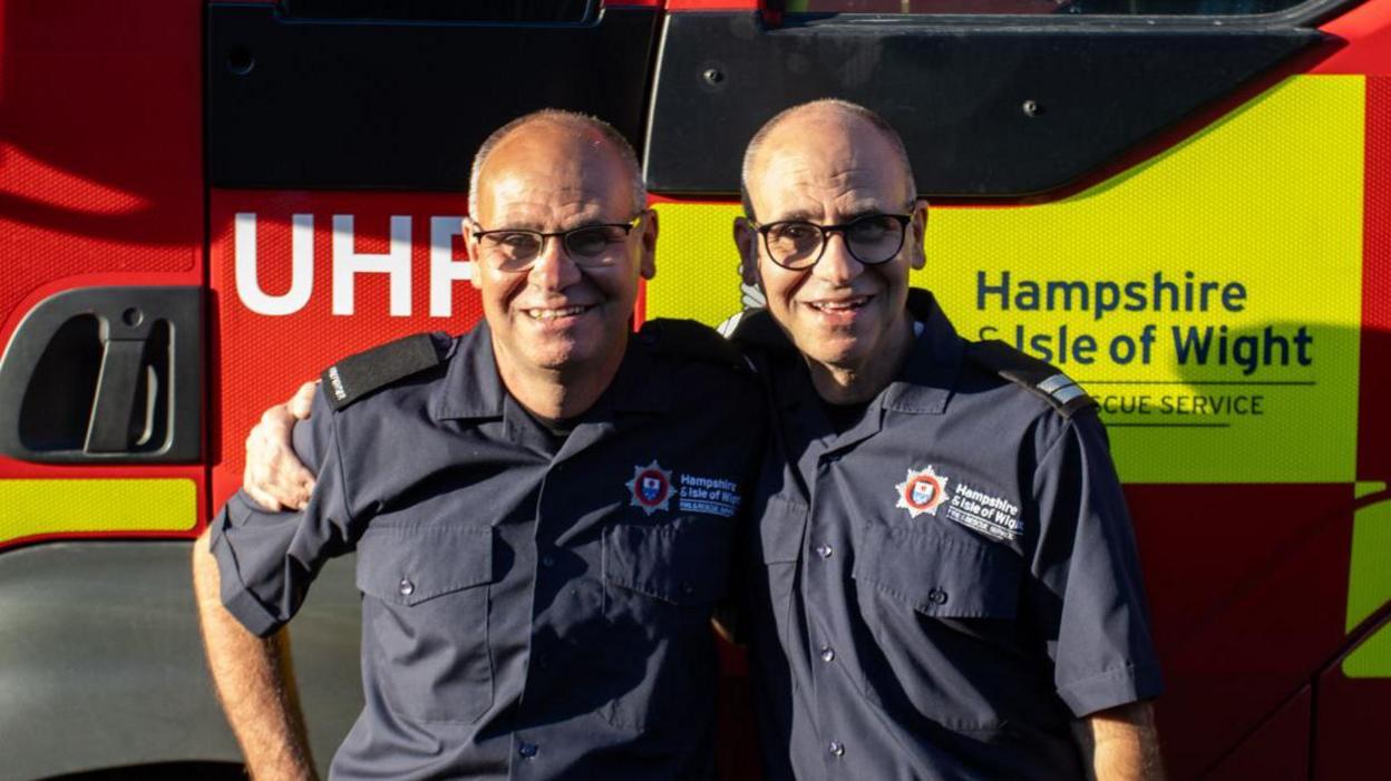 Dave and Paul Minall smiling in front of a Hampshire & Isle of Wight Fire and Rescue Service vehicle. They have their arms around each other's shoulders.
