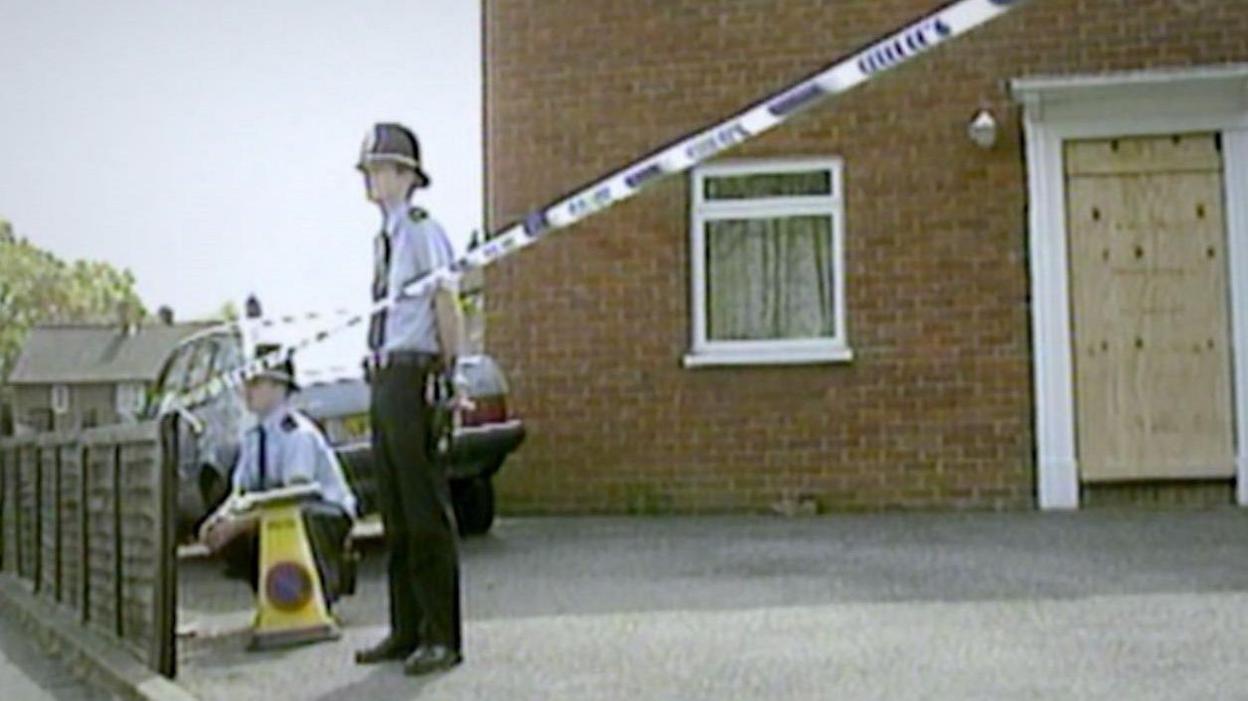 Two police officers outside the home of Karen Reed with police tape running between a fence