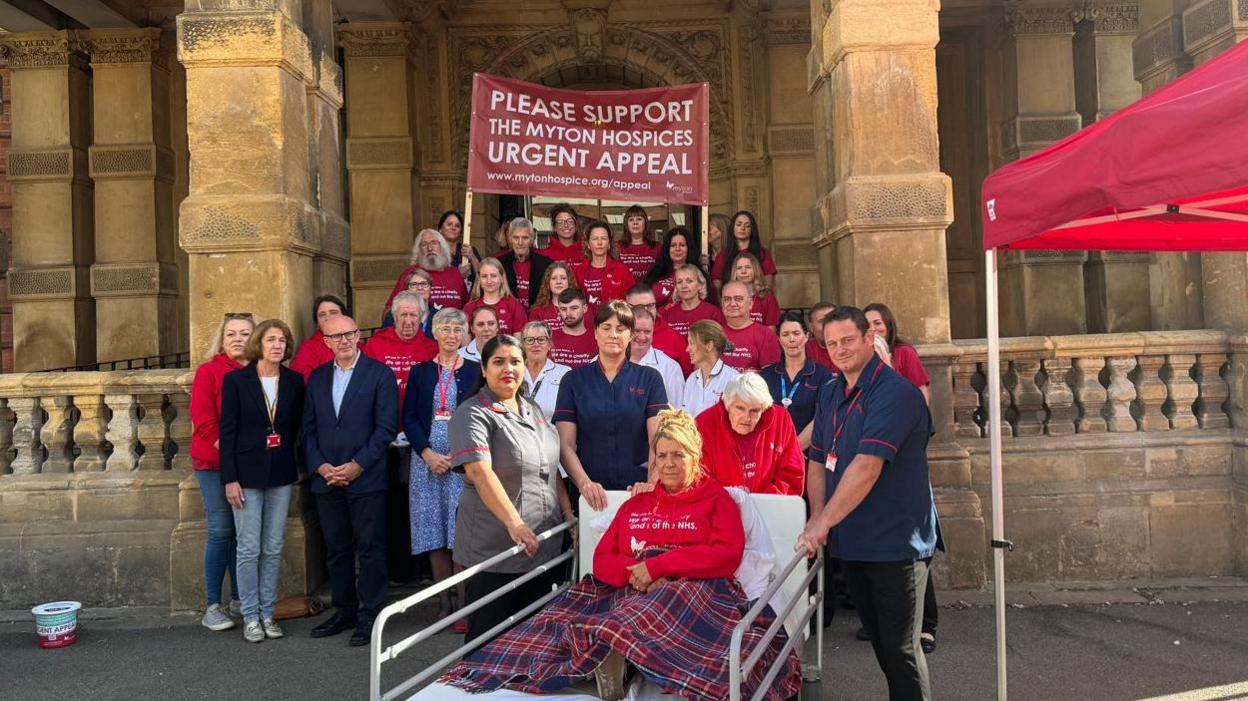 A group of people standing outside Leamington Town Hall. Most of the group are wearing red t-shirts or jumpers with Myton Hospice's logo and some are in nursing uniform. A woman sits in a hospital bed at the front of the group with her legs covered by a chequered blanket.