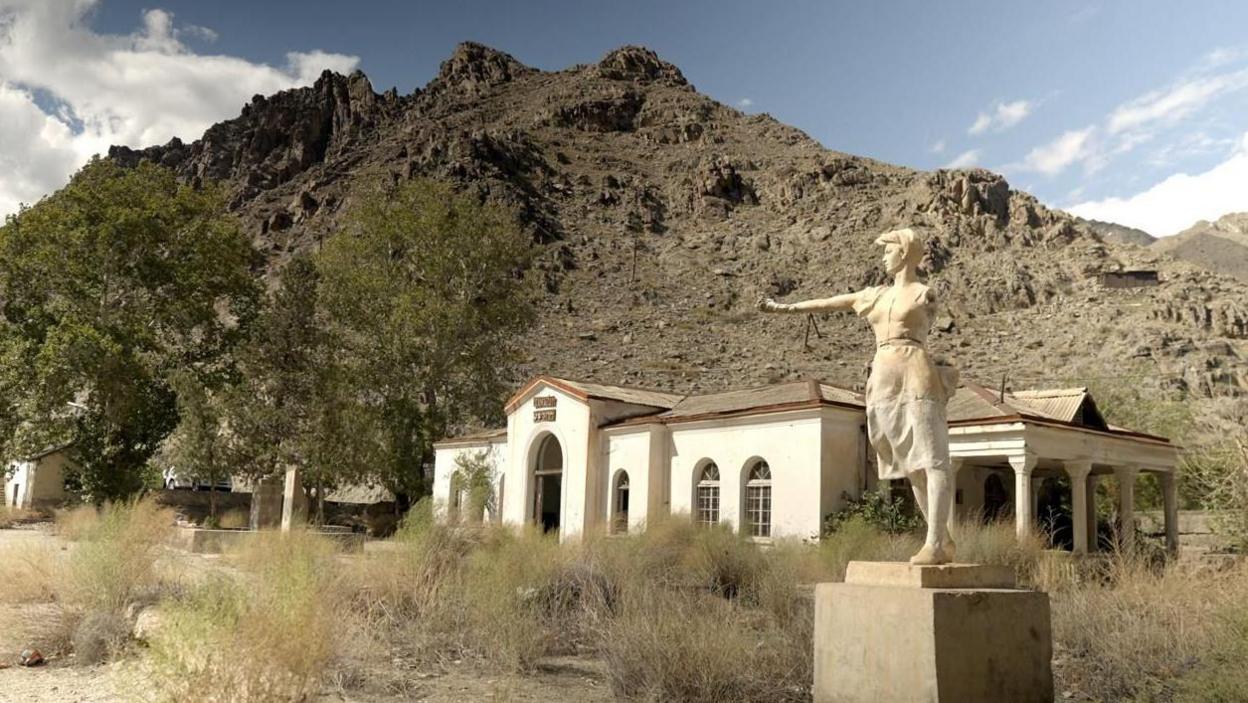 A female sculpture in front of an abandoned railway station with mountains on a backdrop