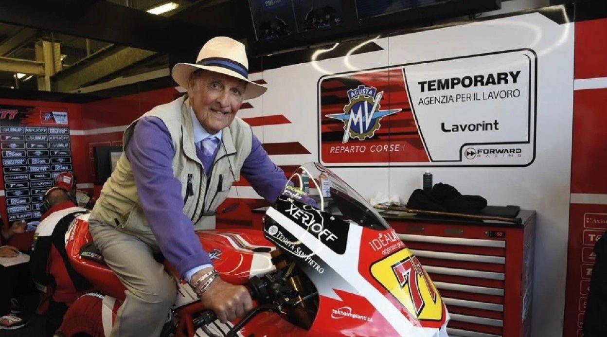 A man wearing a hat, cream gilet and a purple sweater sitting on a red, white and yellow motorbike in a garage