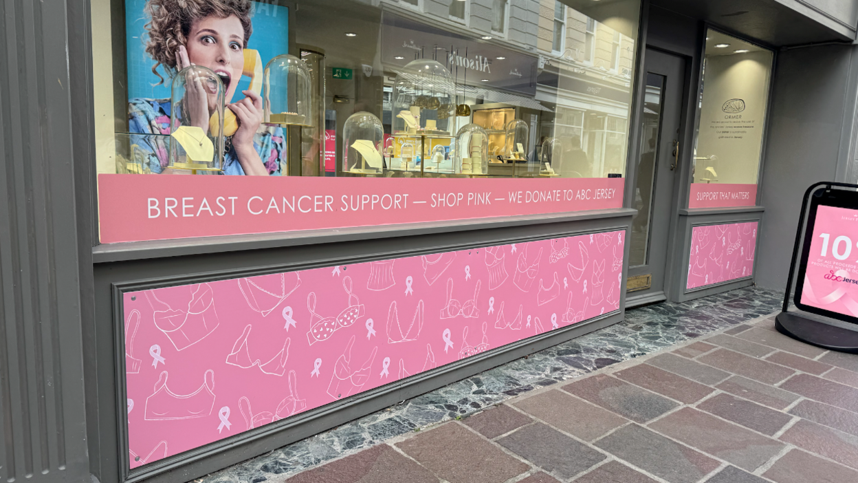 A jewellery shop's window display features a banner below the window with outline drawings of bras and the pink breast cancer ribbon emblem. Above this, stuck to the window itself is a banner which says 'breast cancer support - shop pink - we donate to ABC Jersey'.