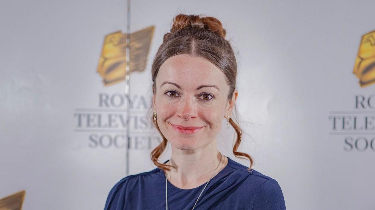 A smiling Sarah Leigh looking directly at the camera, wearing a blue dress and silver necklace. She is standing in front of a poster wall that reads "Royal Television Society"