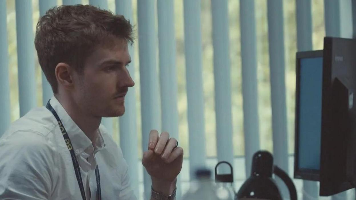 A man wearing a white shirt with short hair is sat at a desk and looking at a computer screen