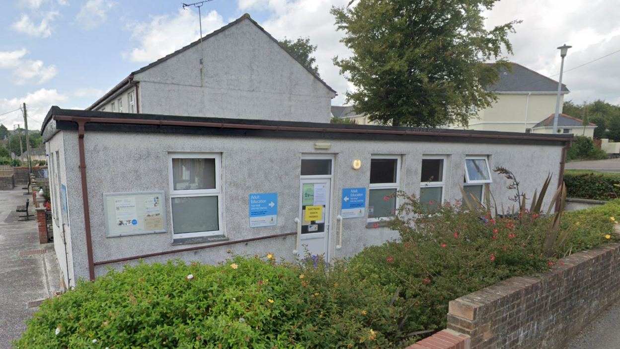 The adult education centre in St Austell is a single storey grey building with shrubs and a red brick wall in front of it. There are sign pointing to the front door that read 'Adult education centre'.