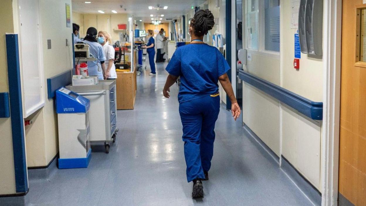 A nurse in blue scrubs is walking along a hospital corridor with her back to the camera. There are doors along the corridor and there is a hub beyond the nurse where other nurses are talking to each other. 