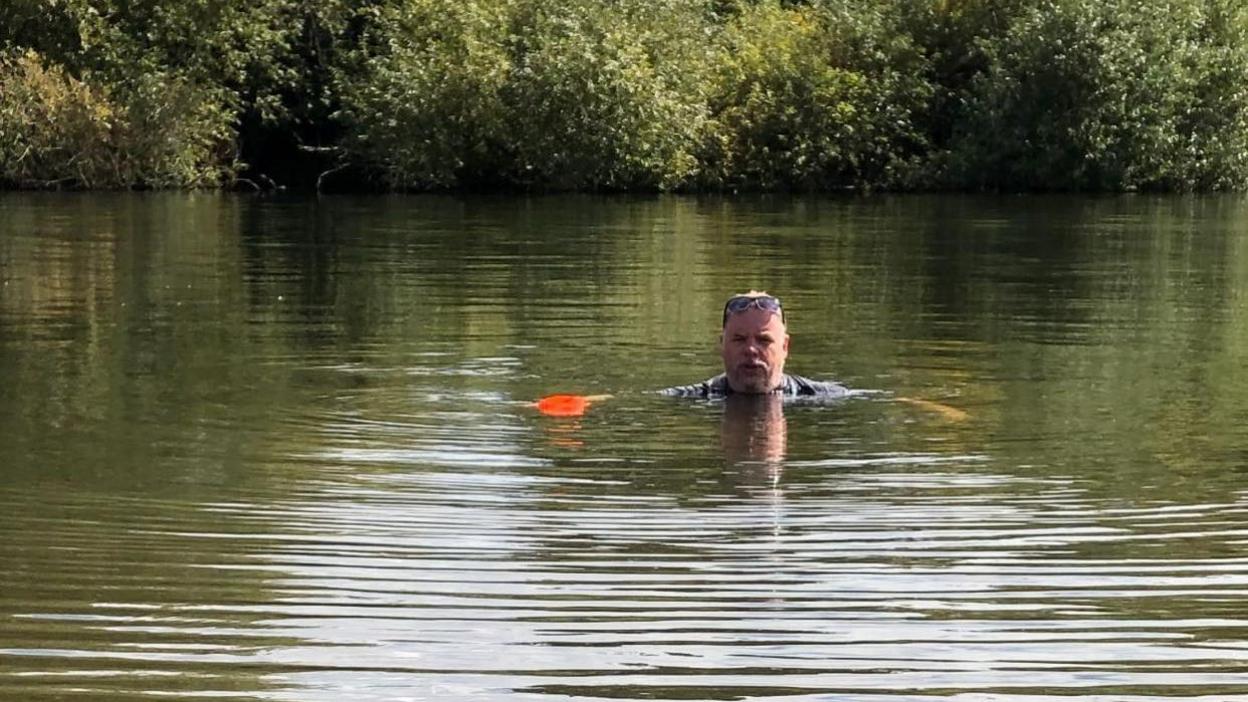 A man swims in a dark green river towards the camera with bushes in the background.