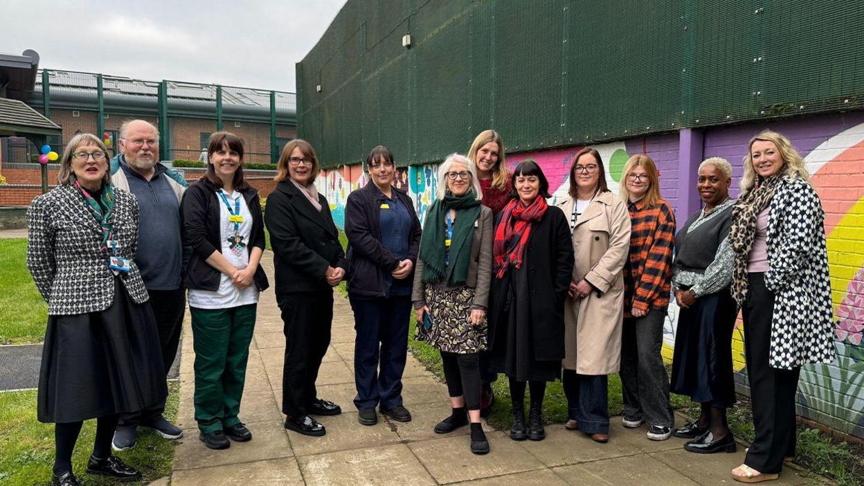 A group of people stand in front of a colourful mural, with a dark green fence in the background.