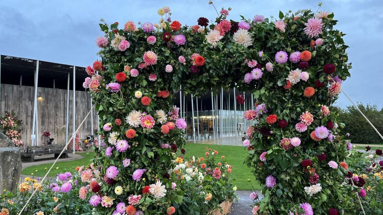 A large archway entrance made from leaves and dahlias. They are all the same colour palette of pink, orange, white and purple.