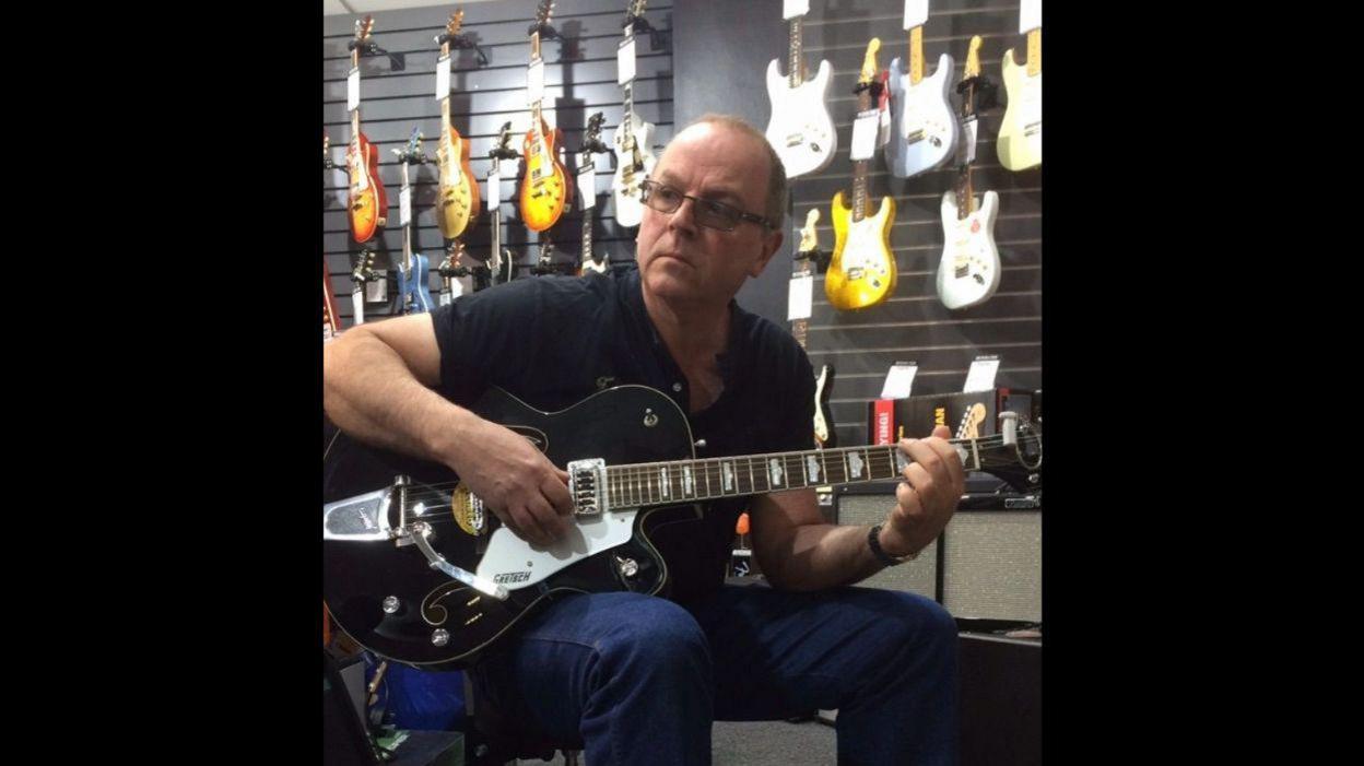 A man in a black t-shirt and jeans is playing a guitar. There is also a wall of guitars behind him.