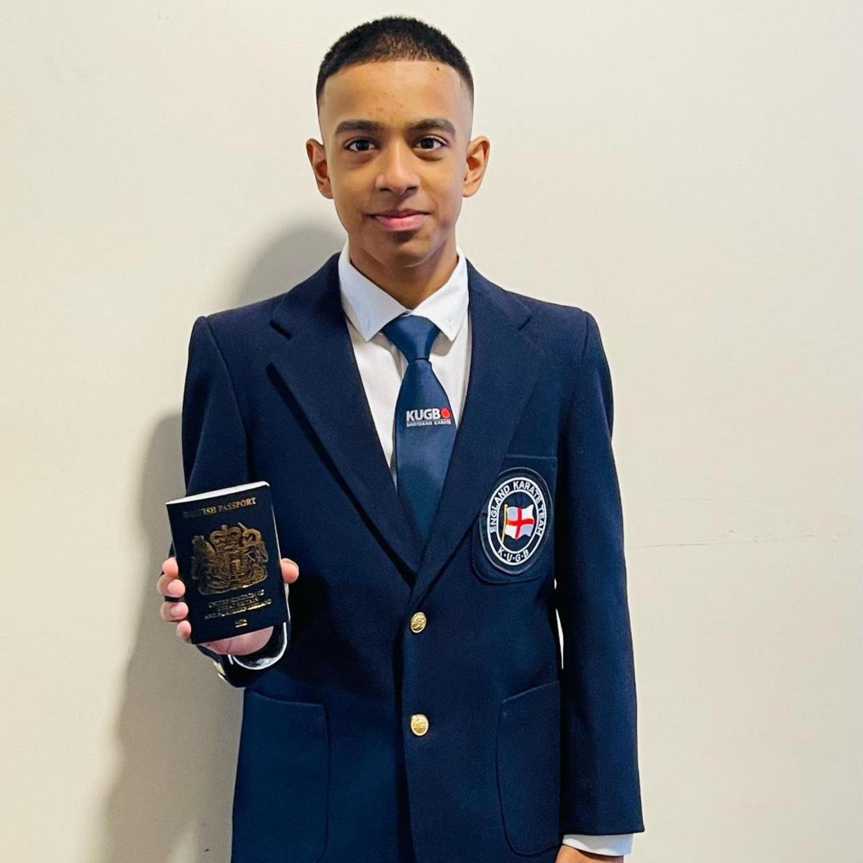 Mayon is holding his passport. He's wearing a navy blazer, embroidered with the England flag on the breast pocket. Around the flag is written "England Karate Team".