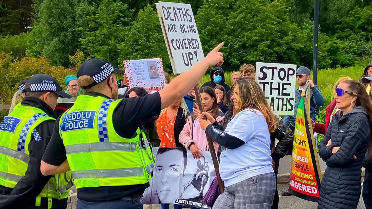 Protestors holding signs saying things like "stop the deaths" stand on a road in front of three police officers