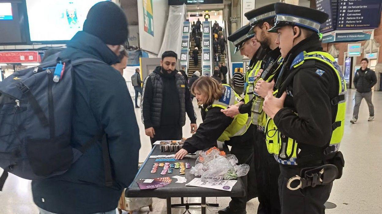Four British Transport Police community officers operate a stall at Victoria Station in London, speaking to three members of the public about White Ribbon Day.