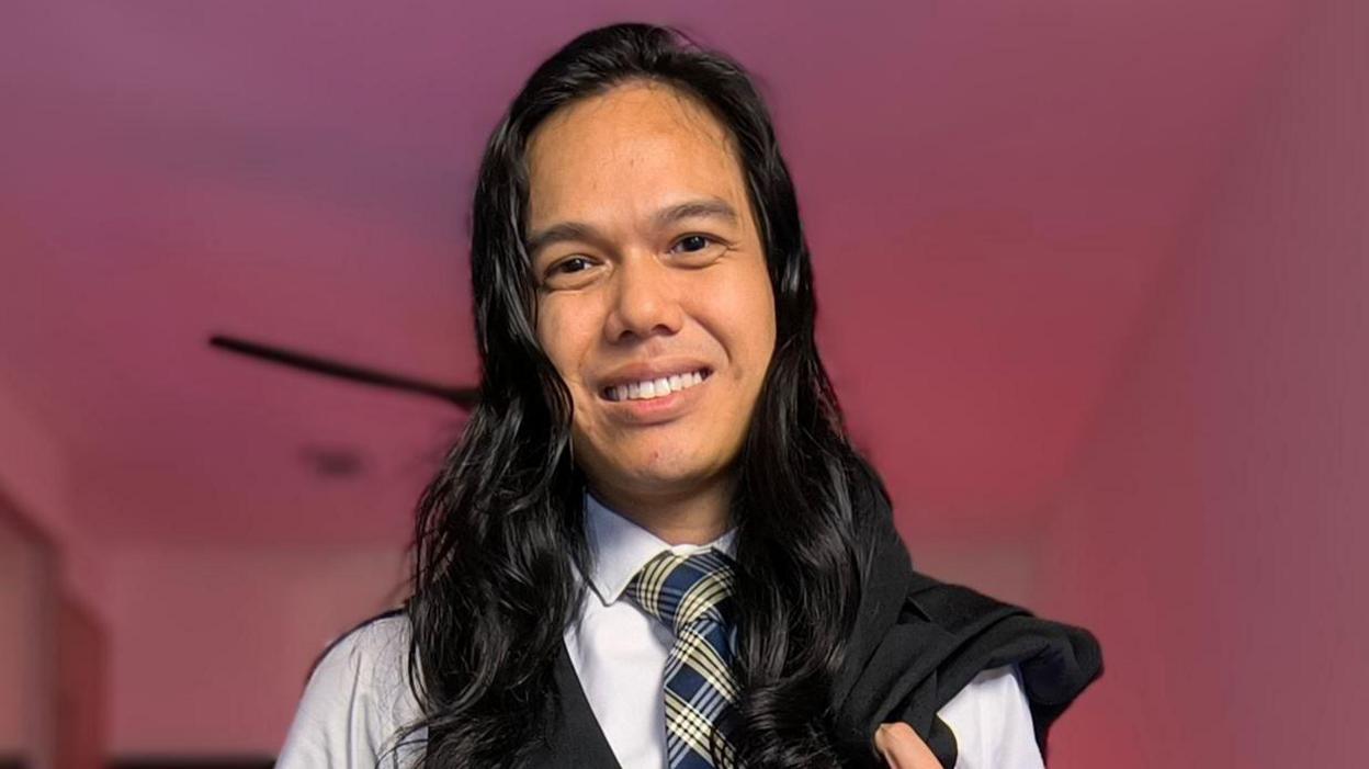 A man with long black hair smiles at the camera, wearing a blue shirt and a checked blue and yellow tie