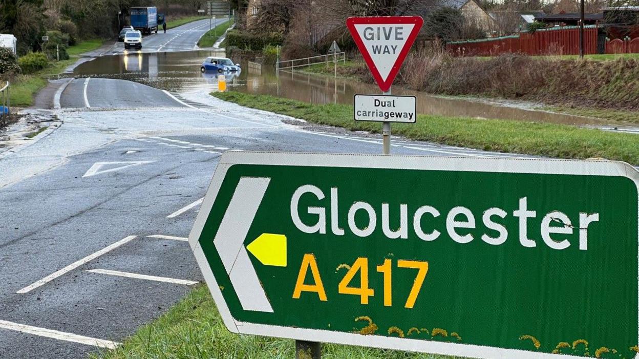 A view of the A417 in Gloucester with road sign in the foreground and a car submerged in floodwater on a road in the background