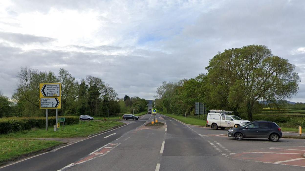 Lock's Garage junction shows the main A465, with roads going off in both directions to Abergavenny and Hay-on-Wye. There are white lines and chevron road markings and give way lines, as well as a bollard. Cars are crossing the junction in the Google Maps image.