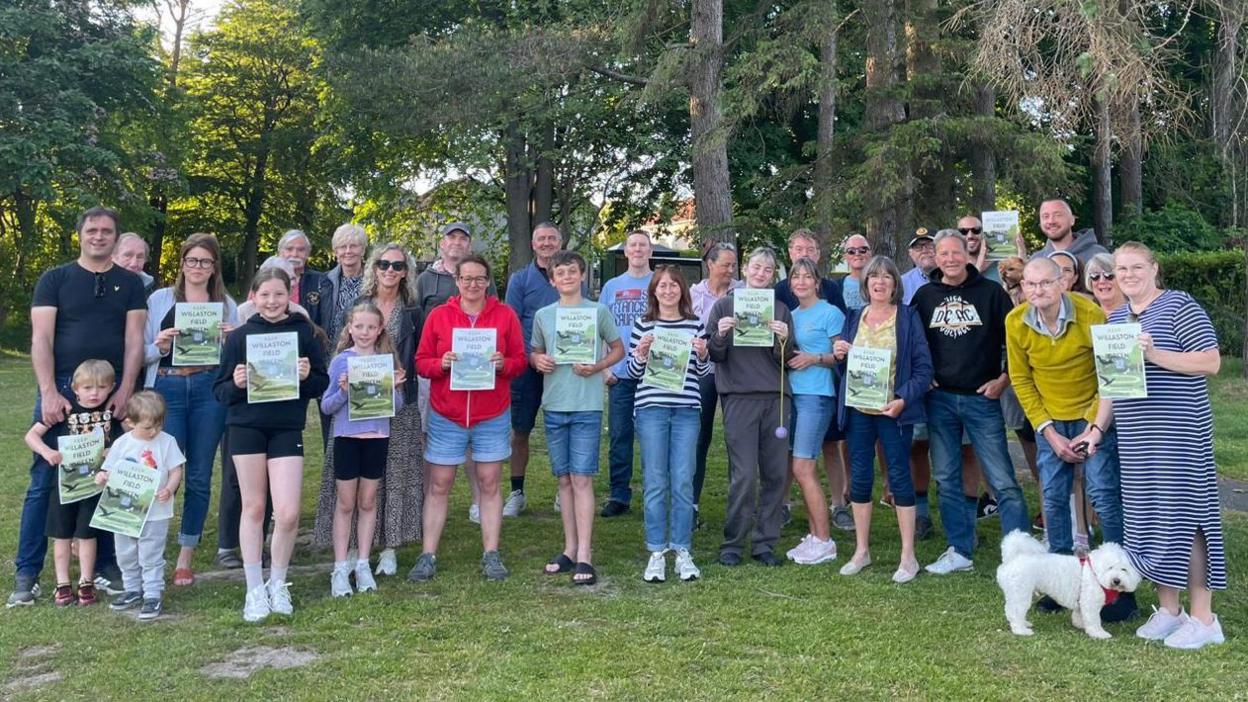 A large group of residents all smiling while holding up posters promoting the group, they stand in the green space, there are children and a white dog.