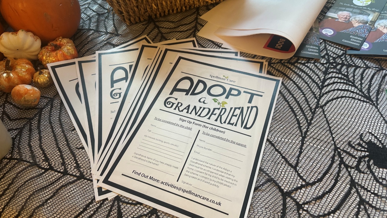 Flyers sit on a table. A table cloth underneath is decorated with spiders webs. The flyers read "adopt a grand-friend". Orange and white pumpkins sit on the table next to the flyers.
