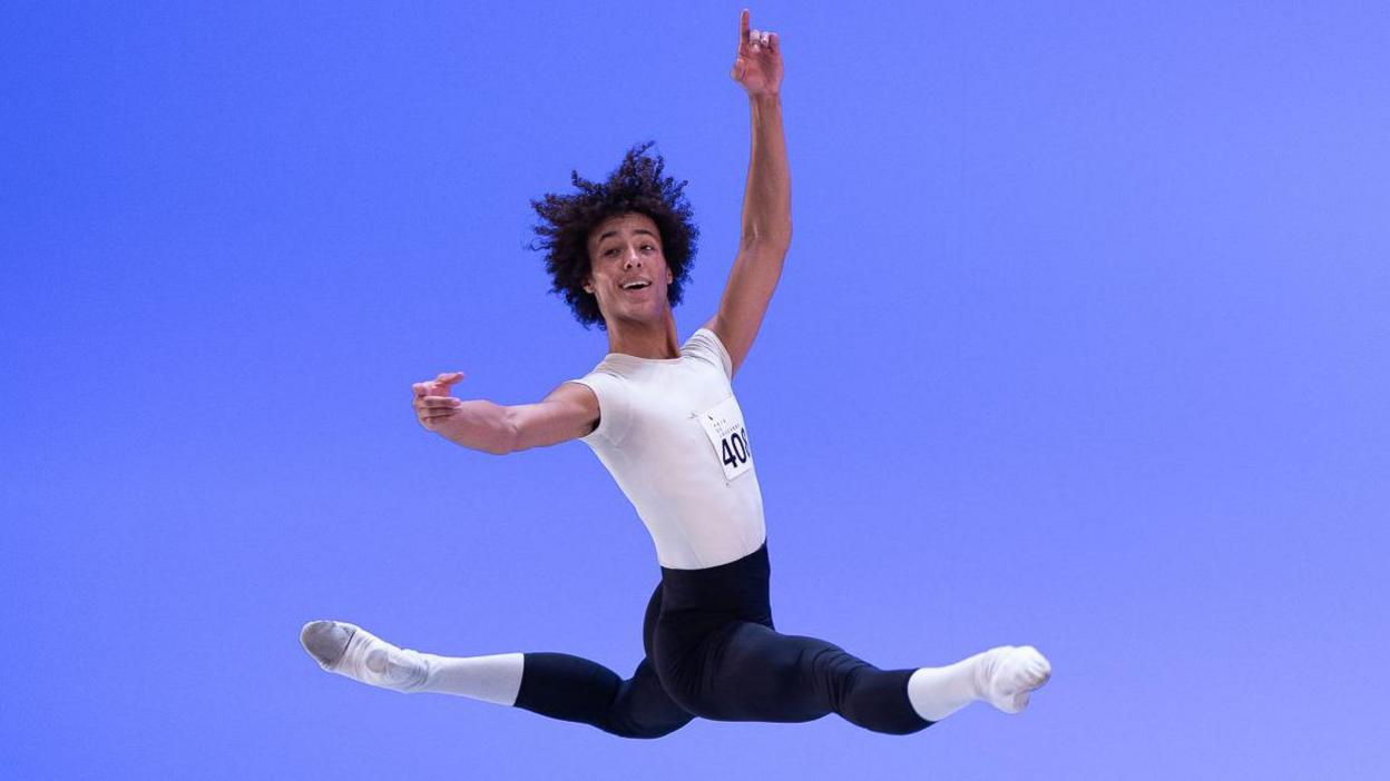 Jakob in full dance position, in the air, with his arms up and legs outstretched. He is wearing a white top with a number on it, black leggings, white socks and white dance shoes. He has short curly hair. 