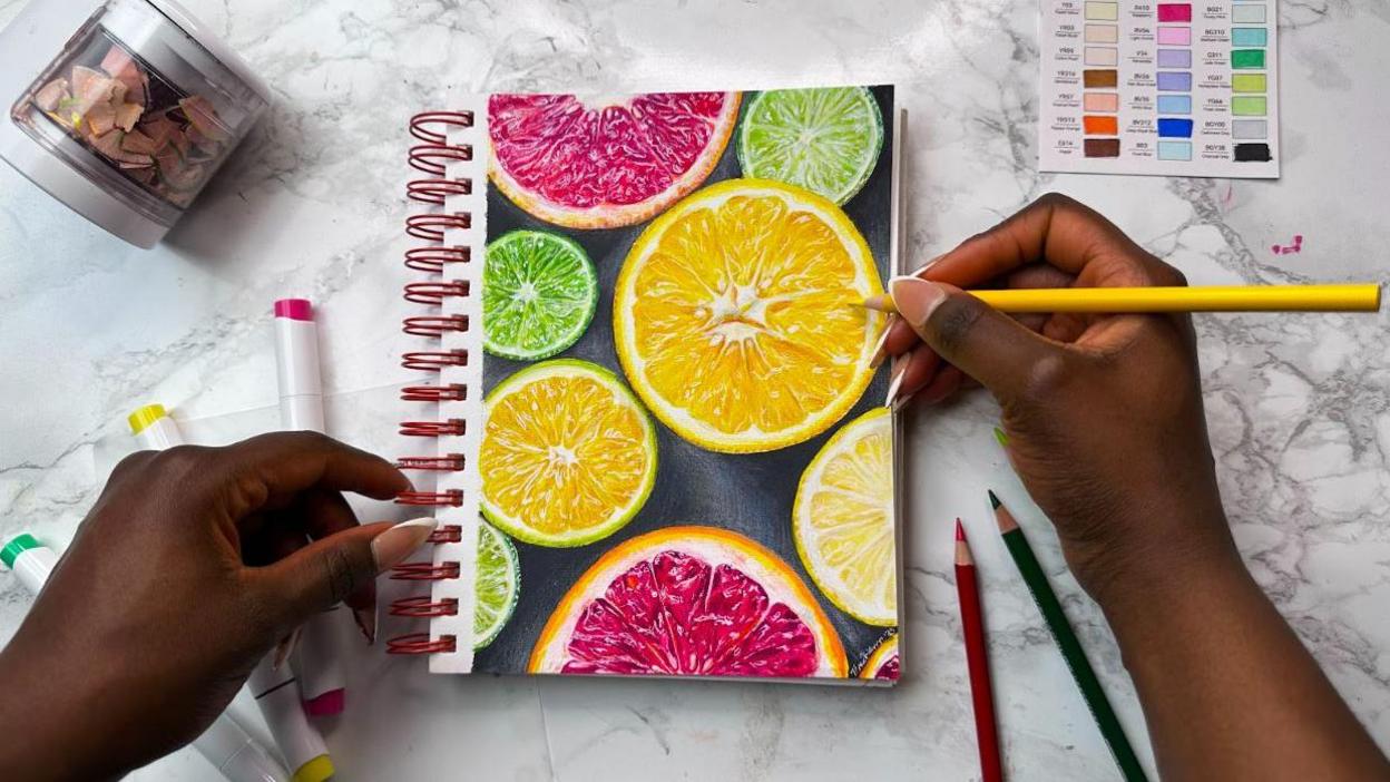 A woman's hands holding a coloured pencil with a drawing of slices of citrus fruit on a notepad in front of her on a white marble surface, with other coloured pencils lying beside it