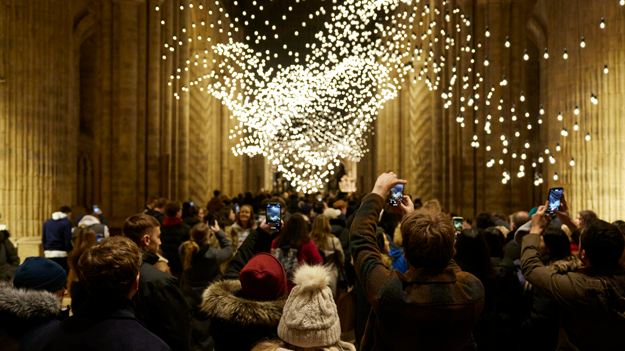 Pulse Topology is an installation of about 4,000 yellow light bulbs, suspended inside Durham Cathedral. Each bulb is activated by the heartbeat of the participants. A crowd of people has surrounded the artwork and some people are taking photos on their phones.