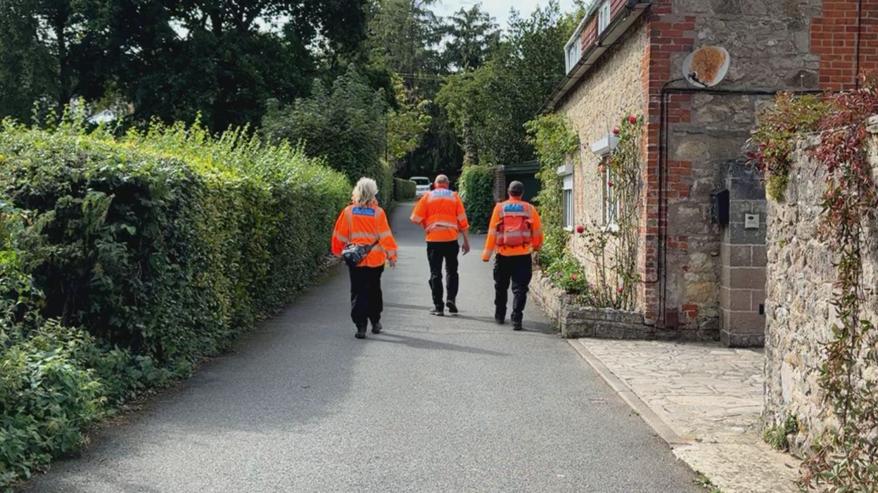 Three people wearing neon orange jackets walking up a country lane. There is a cobblestone house on one side of the road.