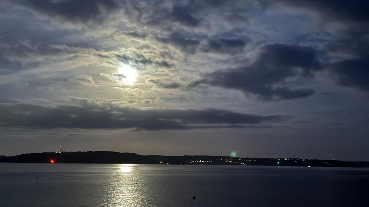 This image captures a peaceful night-time landscape featuring a body of water in the foreground, reflecting the moonlight, a bright moon partially obscured by clouds, casting a soft glow and a distant landmass with scattered lights.