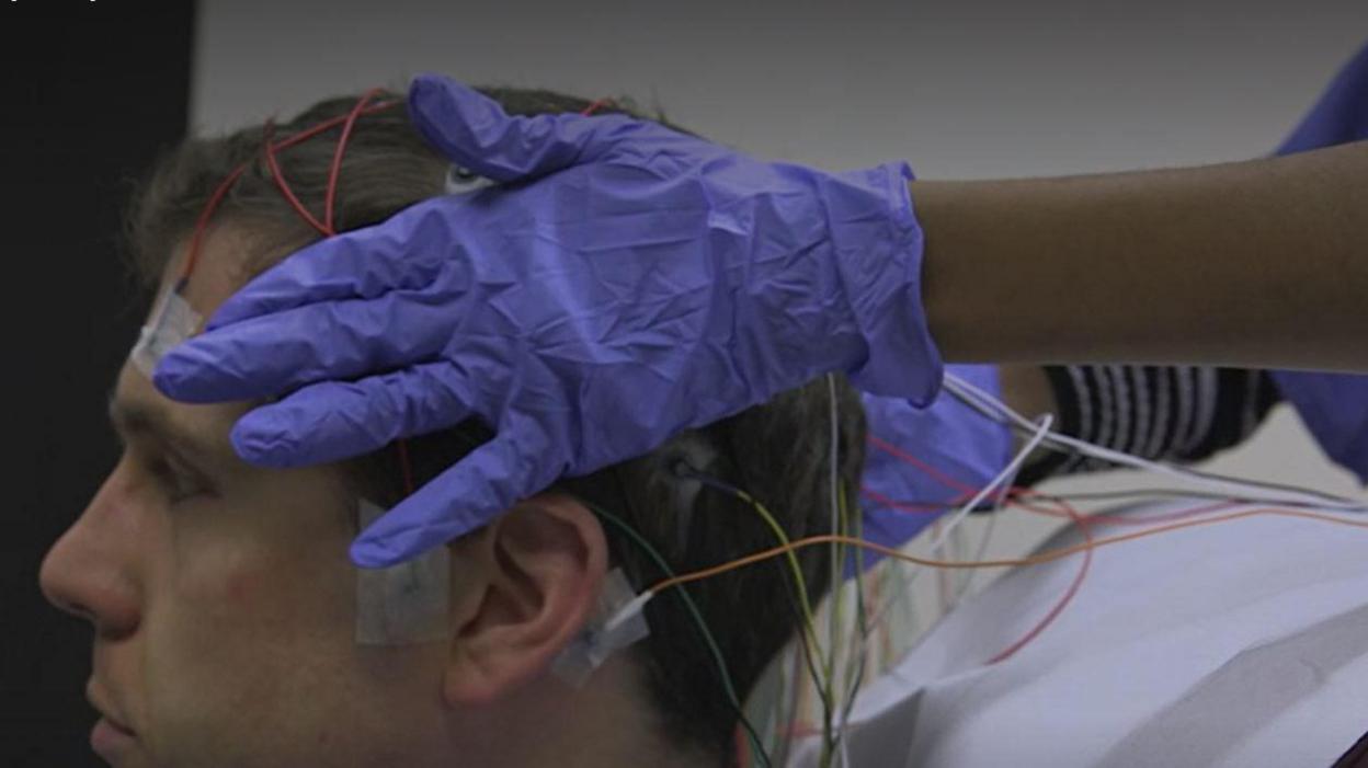 A man with wires stuck to his head and someone with medical gloves holding his head 