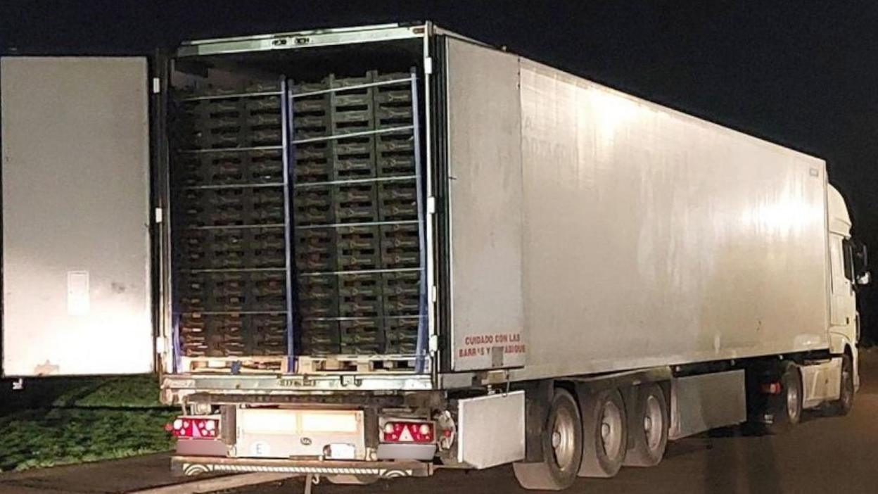 The back of a white lorry has been opened to reveal stacks of produce inside. The lorry has been pulled over at the side of the road and it is night-time.