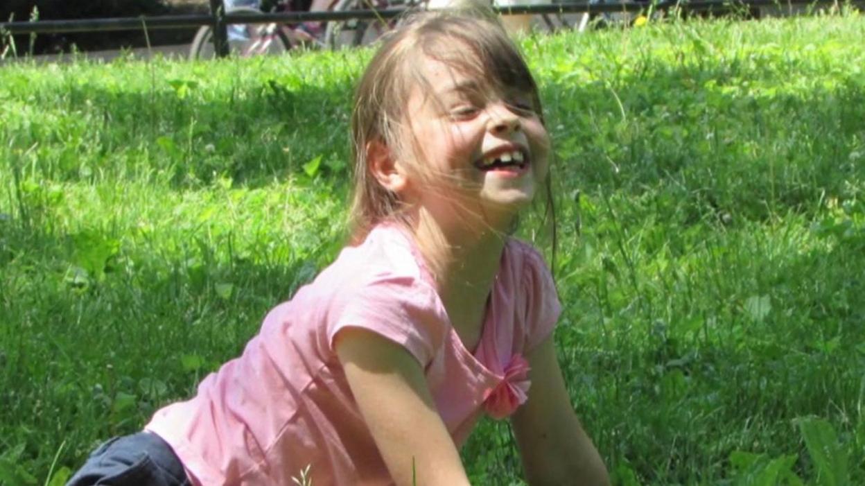 Saffie Roussos is sitting on the grass with a cheeky smile on her face. Her face is looking towards the sky and her hair is blowing in the wind.