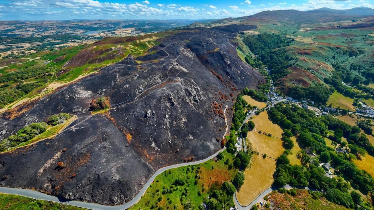 An aerial picture of the Sychnant Pass, with a huge area of hillside burnt and blackened, with the nearby village of Capelulo visible
