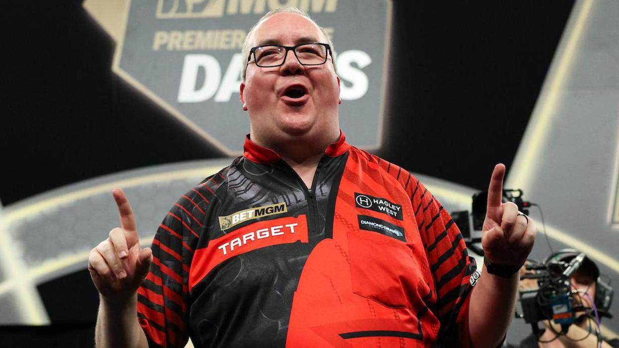 Stephen Bunting celebrates, pointing with both index fingers and pulling a celebratory facial pose as he wears a black and red darts shirt