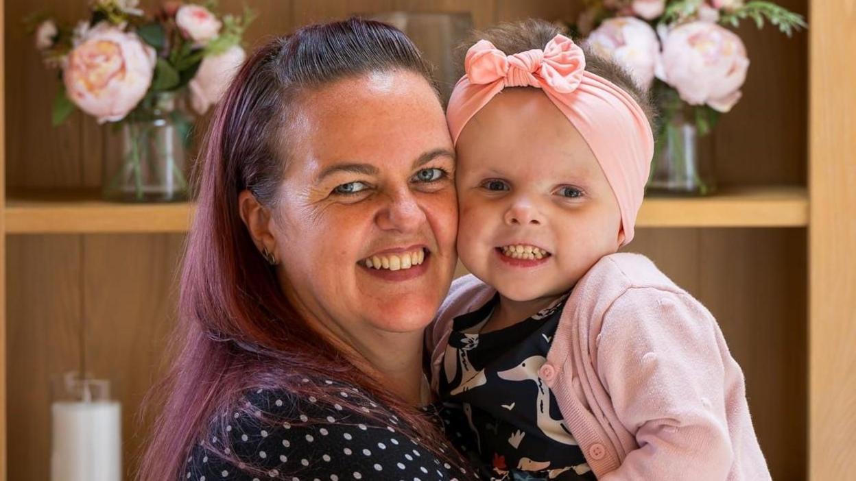 Sally on the left is hugging her daughter Olivia on the right - both are staring directly at the camera and smiling. Olivia is wearing a pink bow on her head and a matching pink cardigan. In the background vases of pink flowers can be seen. Sally has a black and white dress and auburn hair. Photograph was taken as part of Olivia becoming the new face of St Andrew's Hospice.