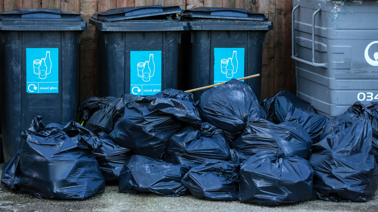 Three black wheelie bins lined up in front of a wooden fence. A pile of black bags lie in the foreground of the photo.