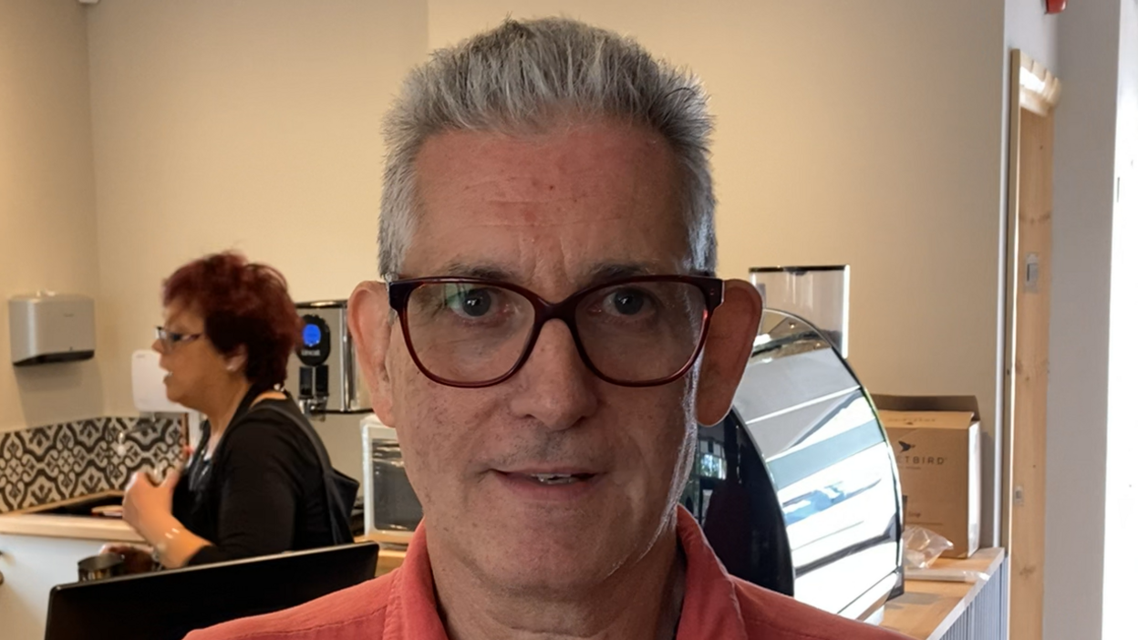 Stuart Hobbs smiles at the camera. He has short grey hair and wears glasses with an orange coloured polo T-shirt. A woman with short red hair stands behind him looking to the left and talking to someone out of frame.