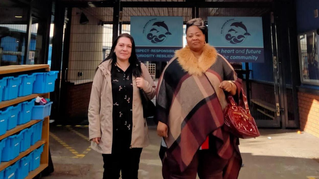 Reneta Atanasova standing next to a friend in front of the Dolphin School. She is wearing a beige coat over a black shirt.