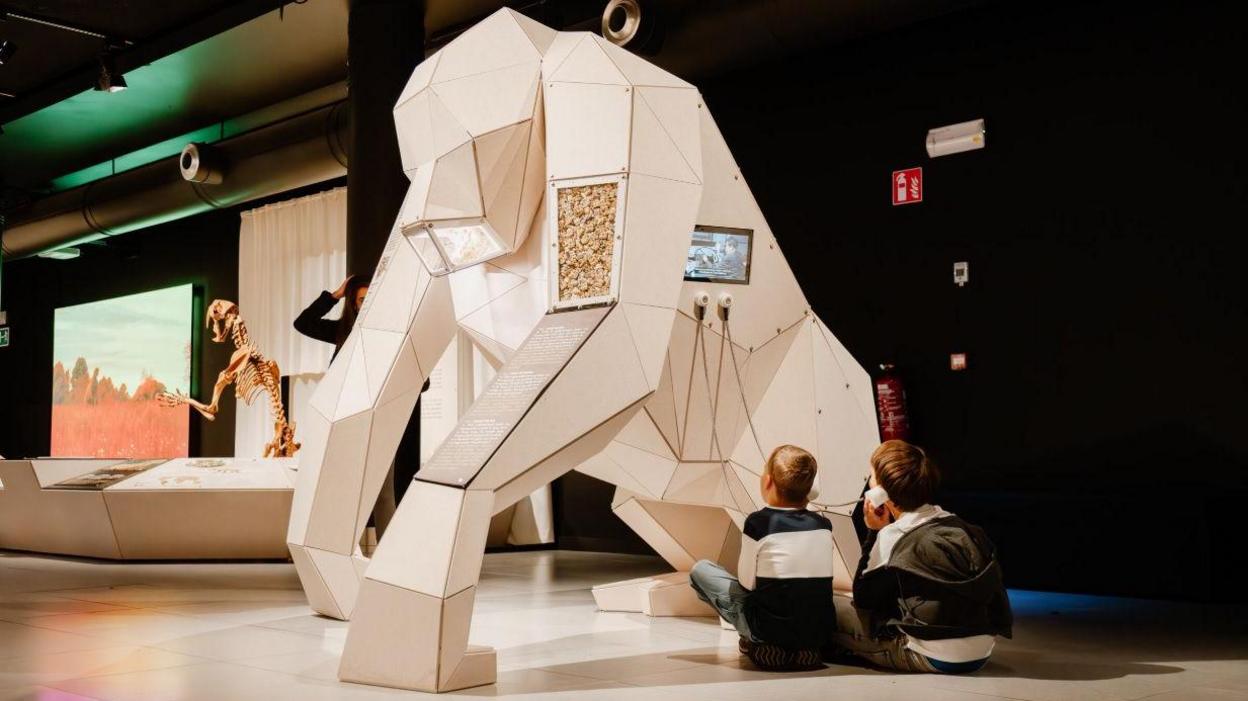 Two children sit in front of a huge model of an ancient primate at the GIANTS Nomad Exhibition.