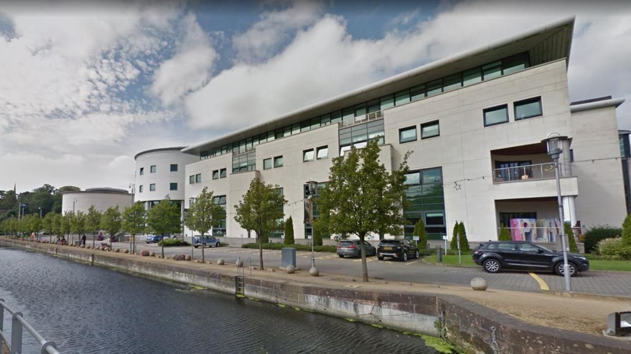 A white building which is the headquarters for Lisburn and Castlereagh Council.
It stand four storeys tall with a flight roof, and rounded section at one end.
It is beside a canal with a number of trees planted in front of it.